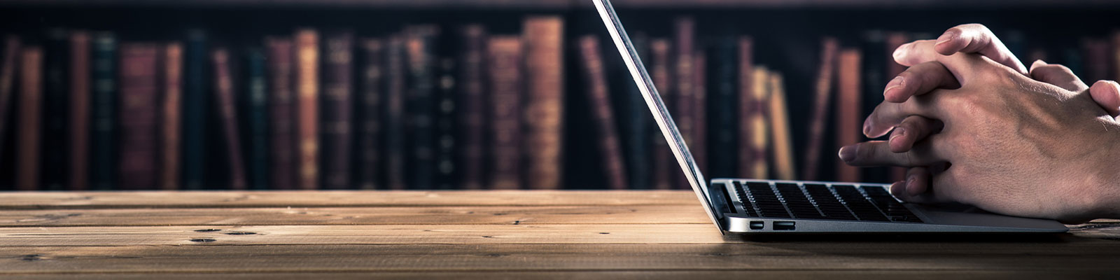hero_library_laptop-stacks_1600x400.jpg open laptop on table in front of shelves of books