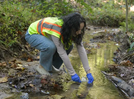 A student bending down to collect a water sample from a local Gwinnett County stream.
