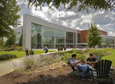 View of the library in background with students sitting in chairs under trees and walking on sidewalk