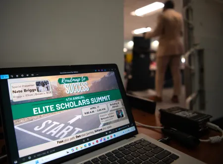 A computer placed on a desk showing "Elite Scholars Summit - Roadmap to Success" graphic on screen