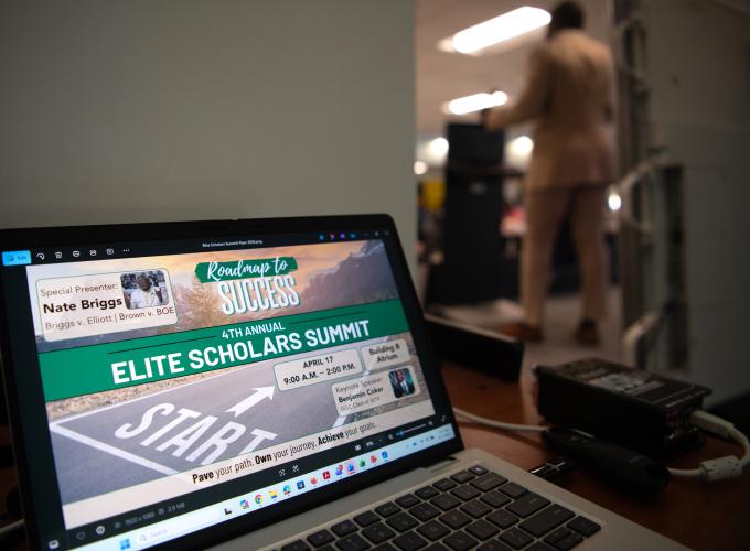 A computer placed on a desk showing "Elite Scholars Summit - Roadmap to Success" graphic on screen
