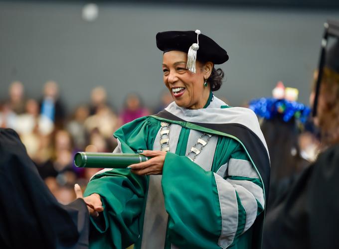 President Jann L. Joseph dressed in commencement cap and gown handing a diploma tube to a graduate during the fall 2025 commencement ceremony