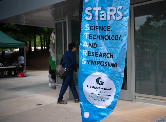 Photo of a light blue background flag banner; "STaRS" written in white letters at tops and Science, Technology, and Research Symposium written horizontally; GGC School of Science and Technology logo at bottom