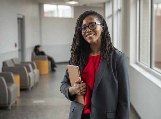 A woman stands in a hall holding a notebook and smiling at a camera; she is wearing glasses, a gray suit and red blouse