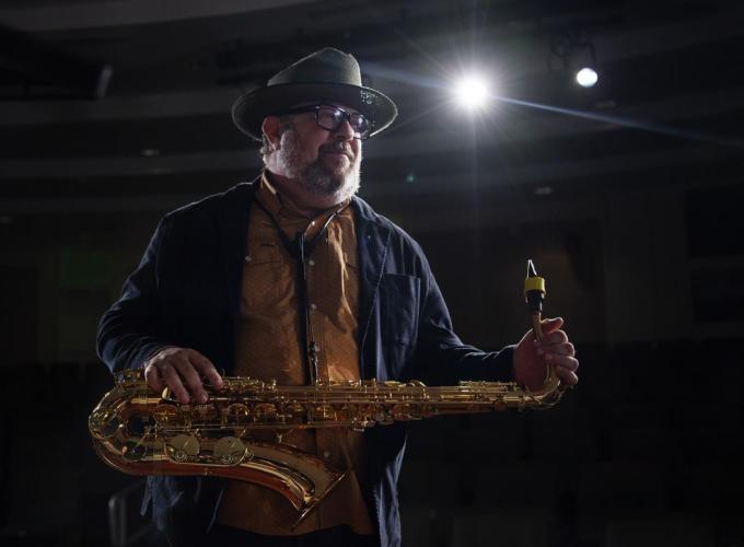 Dr. Marc Gilley on a stage in a dark room with a spotlight shining on him while posing with a saxaphone