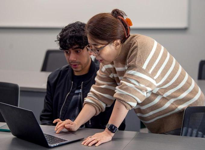 A male student talks with a female professor while looking at a laptop in a classroom.