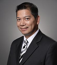 Dr. Binh Tran wearing a black jacket, white shirt and black/white tie and smiling at the camera