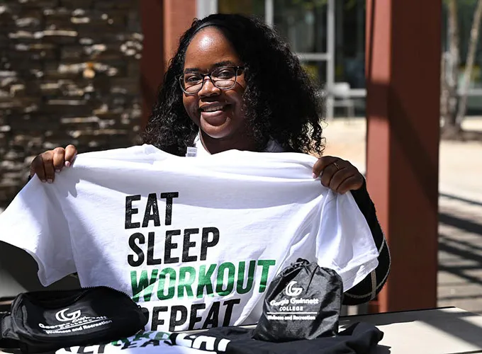Student seated outdoors and holding up a t-shirt that reads eat, sleep, workout, repeat