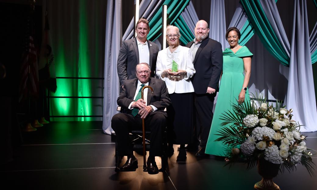 Five people posing for cameras on a stage with gray and green decorative curtains behind them and a live flower arrangement on the right side of the state 