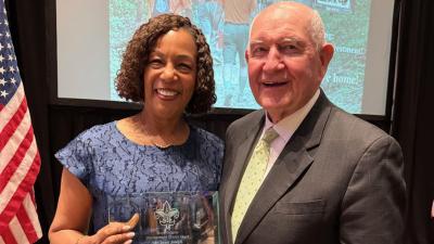 President Jann Joseph standing with a group of Georgia Gwinnett Colelge faculty, staff and Student Government Association president in a large room with two American flags behind them; She is holding the Scott Hudgis Distinguished Citizen Award with Dean Angie Jasper