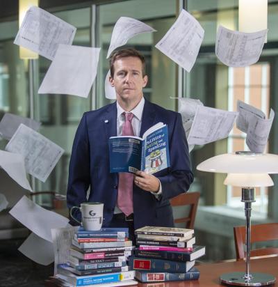 A man standing at a table filled with books; tax papers are floating around him in the air