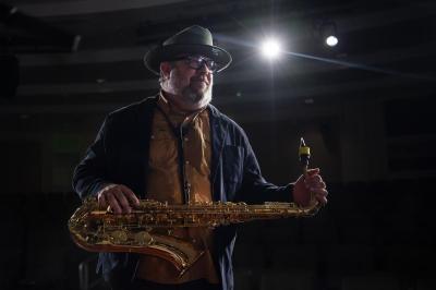 Dr. Marc Gilley on a stage in a dark room with a spotlight shining on him while posing with a saxaphone
